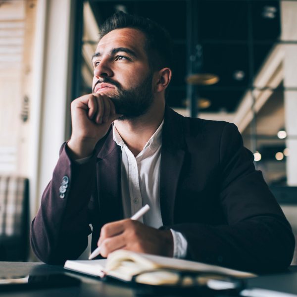Dreamy caucasian bearded business owner looking away thinking about future working in his office, pensive formally dressed male entrepreneur pondering on successful strategy making notes in notepad